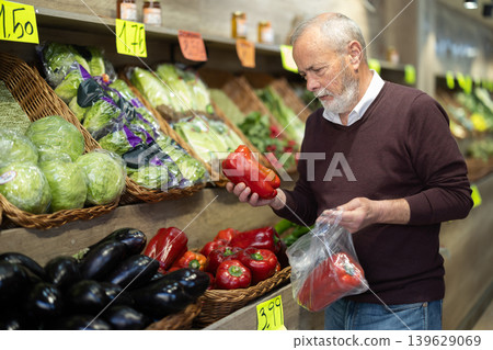 Elderly man stands in the vegetable section of a supermarket and chooses red bell 139629069