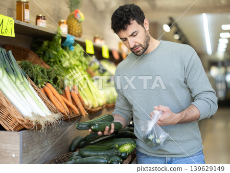 man stands in the vegetable section of a supermarket and chooses zucchini man stands in the vegetable section of a supermarket and chooses zucchini 139629149