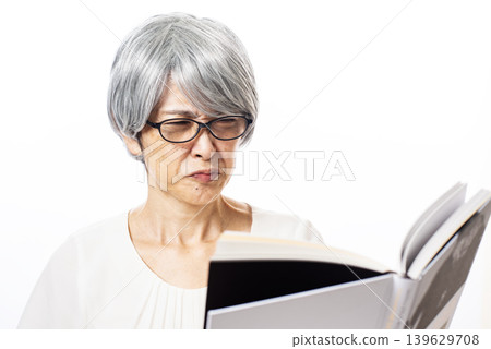 A senior woman with advanced presbyopia reads a book with a frown on her face, wearing black-rimmed reading glasses. A senior woman with advanced presbyopia reads a book with a frown on her face, wearing black-rimmed reading glasses. 139629708