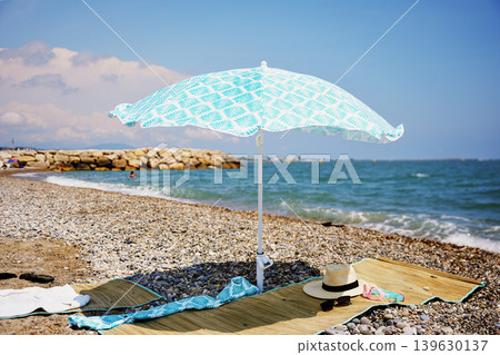 Sunny beach picnic setup with umbrella on pebble shore by the sea Sunny beach picnic setup with umbrella on pebble shore by the sea 139630137