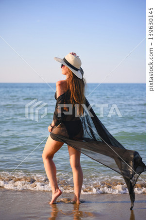 Back view of woman walking on beach in black bikini with sheer cover up Back view of woman walking on beach in black bikini with sheer cover up 139630145