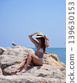 Woman adjusting sun hat while sitting on seaside rocks in summer 139630153