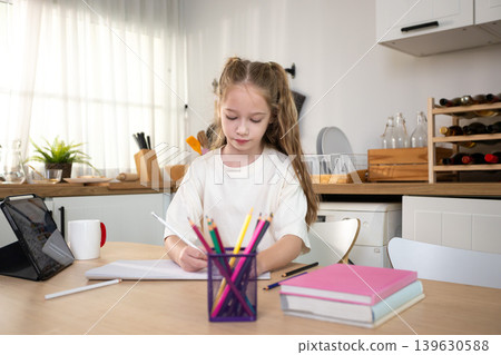 Young girl studying and doing homework at home with digital tablet. Child education, online learning, homeschooling, creativity, and focused learning in a modern kitchen. 139630588