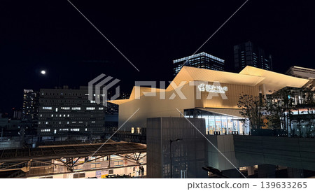 An image of the urban landscape featuring the illuminated Takanawa Gateway Station at night with the moon in the night sky, showcasing its modern architectural design. 139633265