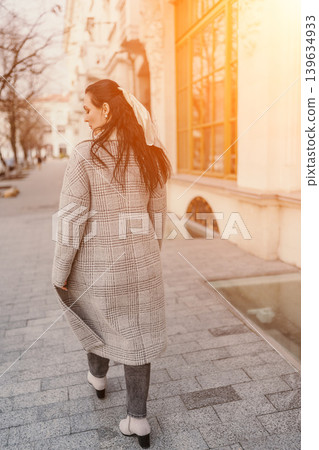 Woman walking sidewalk elegant young brunette in plaid coat strolls down sunlit city street during golden hour with warm sunset glow Woman walking sidewalk elegant young brunette in plaid coat strolls down sunlit city street during golden hour with warm sunset glow 139634933