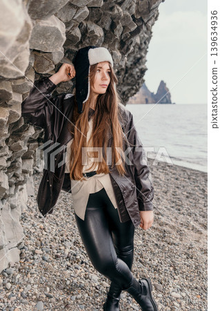 Woman leather jacket ushanka leans against basalt rock formations on a pebble beach during cold coastal travel Woman leather jacket ushanka leans against basalt rock formations on a pebble beach during cold coastal travel 139634936