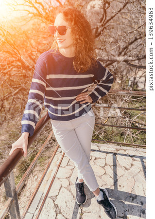 Woman sweater sunset. Young curly haired woman in striped sweater standing on a terrace enjoying golden hour light Woman sweater sunset. Young curly haired woman in striped sweater standing on a terrace enjoying golden hour light 139634963