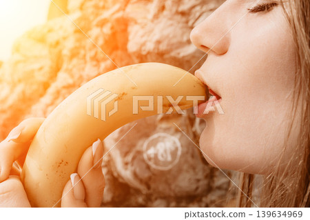 Banana fruit eating woman enjoying healthy yellow snack standing outdoors near rock wall in bright golden hour light 139634969