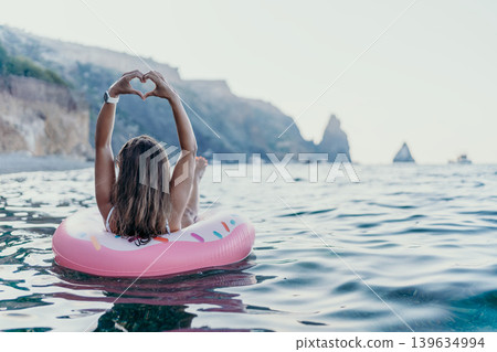 Woman, beach, ocean. Relaxed woman floats on a pink donut tube, forming heart hands in the calm sea with coastal cliffs. Woman, beach, ocean. Relaxed woman floats on a pink donut tube, forming heart hands in the calm sea with coastal cliffs. 139634994