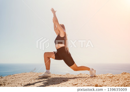 Woman lunge exercise performed on a rocky clifftop overlooking the sea during a sunny outdoor morning fitness workout 139635009