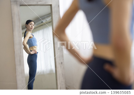 A middle-aged Japanese woman in yoga wear checks her figure in the mirror. A middle-aged Japanese woman in yoga wear checks her figure in the mirror. 139635085