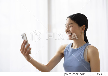一位身穿瑜珈服的日本女子一邊用智慧型手機視訊通話一邊微笑。 139635309