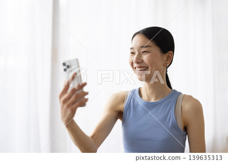 一位身穿瑜珈服的日本女子一邊用智慧型手機視訊通話一邊微笑。 139635313