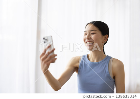 一位身穿瑜珈服的日本女子一邊用智慧型手機視訊通話一邊微笑。 139635314