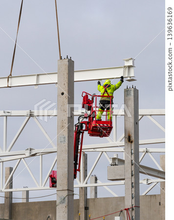 Construction worker in aerial lift aligning steel beam on concrete columns with crane support Construction worker in aerial lift aligning steel beam on concrete columns with crane support 139636169