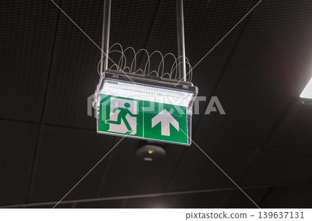 An illuminated green emergency exit sign hangs from a dark ceiling to indicate a safe evacuation route, conveying essential safety, security protocols, and fire prevention in public buildings An illuminated green emergency exit sign hangs from a dark ceiling to indicate a safe evacuation route, conveying essential safety, security protocols, and fire prevention in public buildings 139637131