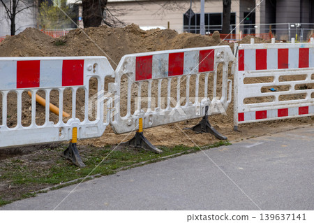 Red and white plastic barriers cordoning off a construction site on a public road, ensuring pedestrian safety and signaling ongoing roadwork maintenance near the sidewalk in an urban setting Red and white plastic barriers cordoning off a construction site on a public road, ensuring pedestrian safety and signaling ongoing roadwork maintenance near the sidewalk in an urban setting 139637141