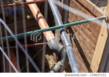 Excavated trench with orange and black pipes reinforced by metal struts for urban infrastructure construction project, highlighting civil engineering development and underground utility systems Excavated trench with orange and black pipes reinforced by metal struts for urban infrastructure construction project, highlighting civil engineering development and underground utility systems 139637151