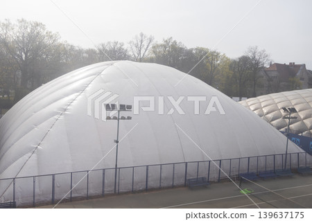 A large white inflatable air dome covers outdoor tennis courts on a misty day, showcasing professional sports infrastructure and seasonal protection for year-round athletic activity enjoyment A large white inflatable air dome covers outdoor tennis courts on a misty day, showcasing professional sports infrastructure and seasonal protection for year-round athletic activity enjoyment 139637175
