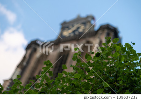 Green leaves in sharp focus with a blurred historical church tower and clock in the background. Scenic urban view of Dordrecht, Netherlands, under a blue sky. Artistic architectural bokeh. Green leaves in sharp focus with a blurred historical church tower and clock in the background. Scenic urban view of Dordrecht, Netherlands, under a blue sky. Artistic architectural bokeh. 139637232