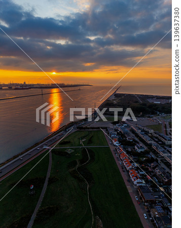 Stunning aerial sunset over a coastal town and industrial port in the Netherlands. Golden light reflects on the water near wind turbines, residential areas, and a scenic park at dusk. 139637380