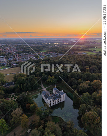 Majestic aerial sunset over a historic moated castle surrounded by dense forest. Golden hour light illuminates the castle architecture and the sprawling European town and countryside beyond. Majestic aerial sunset over a historic moated castle surrounded by dense forest. Golden hour light illuminates the castle architecture and the sprawling European town and countryside beyond. 139637382