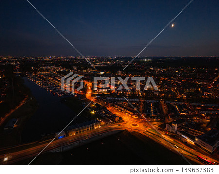 Stunning aerial view of a Dutch city at night under a crescent moon. Warm streetlights illuminate residential neighborhoods, a marina, and roads, creating a vibrant urban landscape at dusk. Stunning aerial view of a Dutch city at night under a crescent moon. Warm streetlights illuminate residential neighborhoods, a marina, and roads, creating a vibrant urban landscape at dusk. 139637383