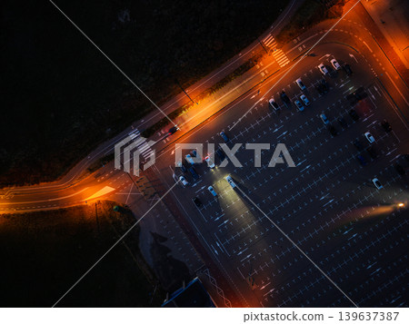 Dramatic aerial top-down view of a parking lot at night with glowing street lamps. Car headlights illuminate the asphalt near a road intersection, creating high contrast patterns in the dark. Dramatic aerial top-down view of a parking lot at night with glowing street lamps. Car headlights illuminate the asphalt near a road intersection, creating high contrast patterns in the dark. 139637387