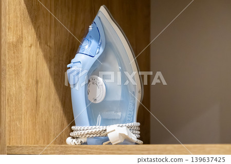 Modern blue steam iron with a coiled cord stored on a wooden shelf, representing home appliance organization, domestic laundry routine, and efficient housekeeping maintenance tasks Modern blue steam iron with a coiled cord stored on a wooden shelf, representing home appliance organization, domestic laundry routine, and efficient housekeeping maintenance tasks 139637425