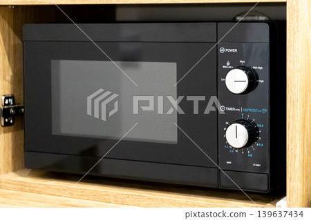 Black microwave oven installed in a wooden kitchen cabinet, featuring manual dial controls for power and timer settings in a modern domestic cooking and food preparation environment Black microwave oven installed in a wooden kitchen cabinet, featuring manual dial controls for power and timer settings in a modern domestic cooking and food preparation environment 139637434