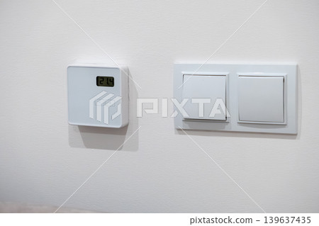 Digital wall thermostat with light switch on white surface, representing modern climate control technology and energy efficient home automation for comfortable living environments 139637435