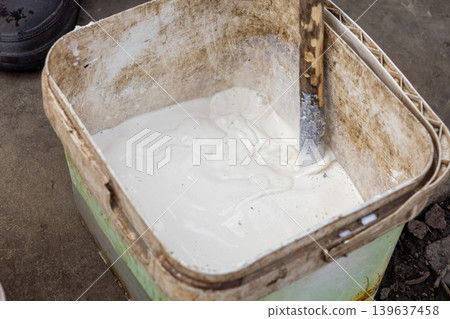 White construction paint being mixed in a weathered plastic bucket with a wooden stick, symbolizing professional renovation, home improvement, DIY repair work, and maintenance tasks 139637458