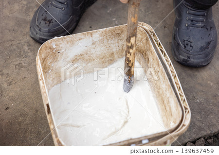 A professional construction worker mixes white paint in a plastic bucket with a wooden stick to prepare for a home renovation project and detailed interior design improvement task 139637459