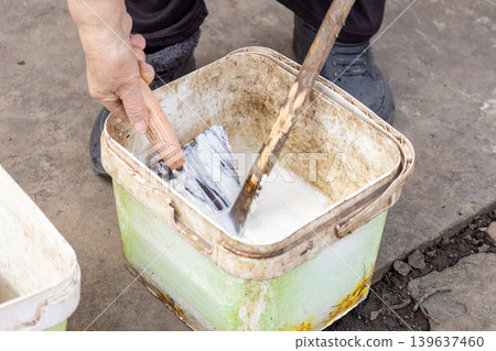 Person mixing white lime mortar in a plastic bucket using a metal trowel and wooden stick, representing preparation of materials for masonry work, plastering, or renovation task Person mixing white lime mortar in a plastic bucket using a metal trowel and wooden stick, representing preparation of materials for masonry work, plastering, or renovation task 139637460