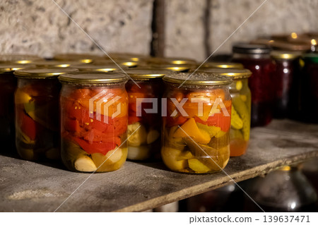 Glass jars filled with pickled peppers and garlic on a wooden shelf in a dark cellar, representing home food preservation and traditional preparation of seasonal vegetable harvest Glass jars filled with pickled peppers and garlic on a wooden shelf in a dark cellar, representing home food preservation and traditional preparation of seasonal vegetable harvest 139637471