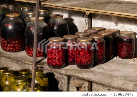 Glass jars with preserved cherries and pickles on wooden cellar shelves illustrate traditional food preservation, rustic pantry organization, and a sustainable rural home lifestyle Glass jars with preserved cherries and pickles on wooden cellar shelves illustrate traditional food preservation, rustic pantry organization, and a sustainable rural home lifestyle 139637472