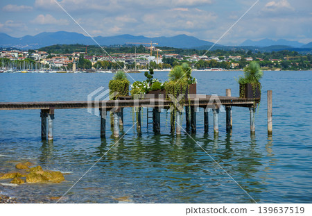 Wooden pier with green plants and flowers over clear water, scenic lakeside view with distant town and mountains under bright summer sky. Wooden pier with green plants and flowers over clear water, scenic lakeside view with distant town and mountains under bright summer sky. 139637519