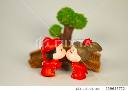 Adorable miniature bride and groom figurines in red traditional clothing sharing a romantic moment beside a decorative bridge and tree background. Adorable miniature bride and groom figurines in red traditional clothing sharing a romantic moment beside a decorative bridge and tree background. 139637752