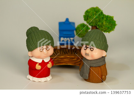 Charming elderly miniature figurines wearing knitted winter outfits beside a small mailbox and tree scene on a clean white background. 139637753