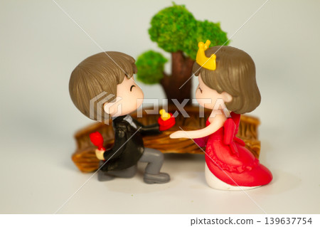 Cute miniature prince and princess figurines in a proposal moment with a crown and ring beside a decorative tree and wooden bridge background. Cute miniature prince and princess figurines in a proposal moment with a crown and ring beside a decorative tree and wooden bridge background. 139637754