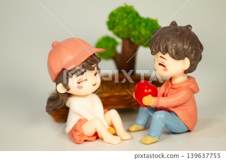 Adorable miniature couple figurines sharing a sweet heart gift moment beside a decorative tree and wooden bridge in a playful romantic setting. Adorable miniature couple figurines sharing a sweet heart gift moment beside a decorative tree and wooden bridge in a playful romantic setting. 139637755