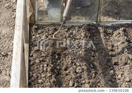 Gardener using a hoe to cultivate soil inside a wooden greenhouse, preparing the garden bed for planting seasonal vegetables and herbs to promote sustainable organic farming and food growth 139638106