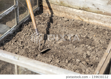 Old hand hoe resting in dark soil inside a wooden garden cold frame, reflecting rural spring preparation and organic vegetable cultivation for sustainable food growth in a backyard setting 139638112
