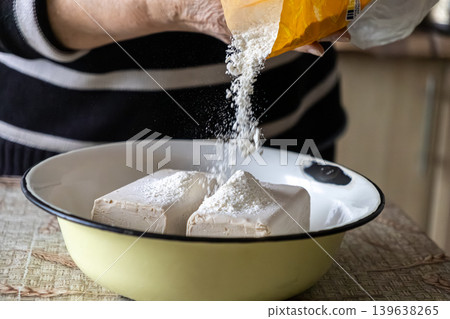 Person pouring wheat flour over fresh yeast blocks in a bowl, capturing the authentic process of preparing homemade dough for artisan bread baking in a rustic domestic kitchen Person pouring wheat flour over fresh yeast blocks in a bowl, capturing the authentic process of preparing homemade dough for artisan bread baking in a rustic domestic kitchen 139638265