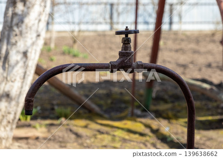 An old rusty metal water tap on a curved pipe in a garden illustrates themes of agricultural neglect, water supply infrastructure, and rustic outdoor plumbing maintenance concepts An old rusty metal water tap on a curved pipe in a garden illustrates themes of agricultural neglect, water supply infrastructure, and rustic outdoor plumbing maintenance concepts 139638662