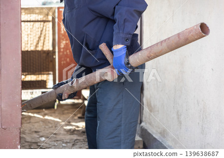 Worker in protective gloves carrying a rusty metal pipe on a construction site, emphasizing labor, manual task, infrastructure maintenance, and professional industrial activity Worker in protective gloves carrying a rusty metal pipe on a construction site, emphasizing labor, manual task, infrastructure maintenance, and professional industrial activity 139638687