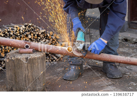 An industrial worker uses a powerful angle grinder to cut through a thick metal pipe, creating bright sparks and flying debris while demonstrating precision skill in construction An industrial worker uses a powerful angle grinder to cut through a thick metal pipe, creating bright sparks and flying debris while demonstrating precision skill in construction 139638717