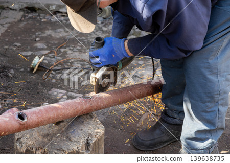 An industrial worker carefully cuts through a thick rusty steel pipe with an angle grinder while bright orange sparks fly across the dark workshop during a professional metal fabrication job 139638735