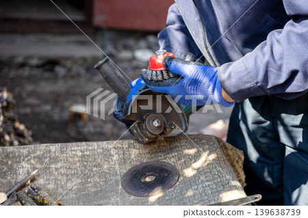 Worker in protective gloves holds an angle grinder with a wire brush attachment for metal cleaning, demonstrating manual labor, home renovation, and industrial maintenance processes outdoors Worker in protective gloves holds an angle grinder with a wire brush attachment for metal cleaning, demonstrating manual labor, home renovation, and industrial maintenance processes outdoors 139638739