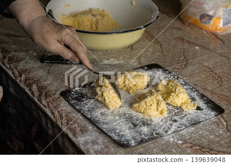 A senior woman shapes traditional syrniki cottage cheese pancakes on a floured wooden board, representing culinary heritage and authentic homemade cooking techniques in a rustic home kitchen A senior woman shapes traditional syrniki cottage cheese pancakes on a floured wooden board, representing culinary heritage and authentic homemade cooking techniques in a rustic home kitchen 139639048